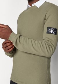 Chemise longue olive texturée à manches longues avec un col rond et des poignets côtelés. Équipée d'un patch de logo noir Calvin Klein Jeans sur la manche.