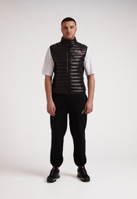 Gilet imbottito nero con motivo trapuntato orizzontale, colletto alto, zip frontale e logo. Indossato sopra una t-shirt bianca e pantaloni sportivi neri.