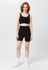Czarny crop top z szerokimi ramiączkami i czarnymi szortami z wysokim stanem. Materiał jest gładki i dopasowany. Model nosi białe sneakersy i skarpetki.