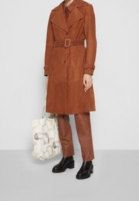 Trench coat de ante en color óxido, con solapas anchas, cinturón y cierre de botones. Combinado con pantalones marrones de cuero sintético y botas con tacón ancho.