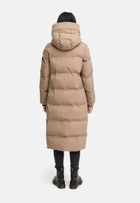 Manteau rembourré long beige avec une grande capuche, conçu avec des sections matelassées. Porté sur un pantalon noir et des bottes noires robustes.