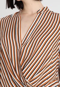 Blouse rayée en orange, blanc et noir. Présente un design croisé avec un col et une texture de tissu lisse. Les rayures verticales dominent le motif.