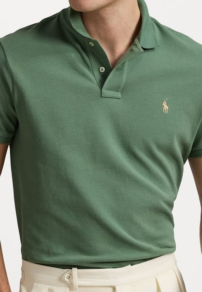 Zaļš polo krekls ar teksturētu audumu, divu pogu aizdari un mazu izšūtu logotipu pelēkā krāsā kreisajā krūtī. Kombinēts ar baltiem biksēm.