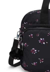 Svart tygväska med ett blommönster i lila och blått. Har ett ficka med dragkedja och en fluffig gorillanyckelring fäst vid remmen.