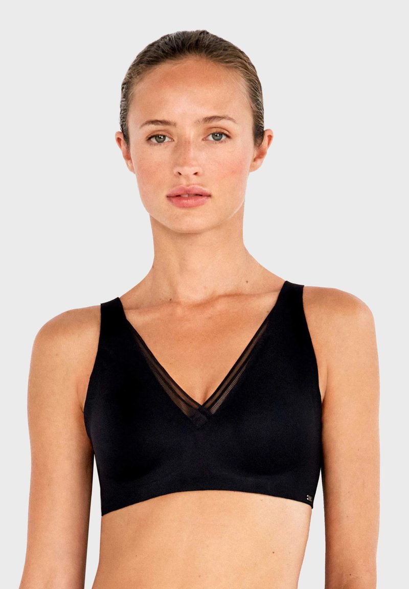 Femme de face, portant un soutien-gorge noir sans armatures avec un empiècement en tulle transparent en forme de V, expression neutre, fond uni gris clair.