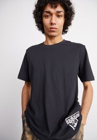 Μαύρο βαμβακερό T-shirt με στρογγυλή λαιμόκοψη, κοντά μανίκια και λευκή ετικέτα με το λογότυπο της Adidas στο αριστερό ισχίο. Απλή ύφανση με λεία υφή.