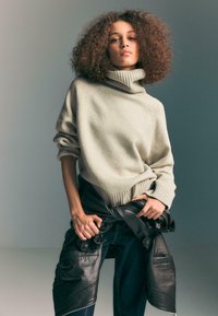 Giovane con capelli ricci che indossa un maglione beige a collo alto e jeans scuri, stringendo una giacca di pelle nera attorno alla vita.