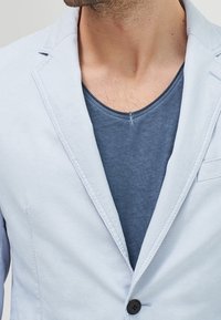Světle modrý blazer s texturovanou tkaninou, jedním černým knoflíkem a špičatými klopami, nošený přes tmavě modré tričko.
