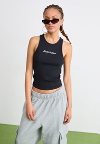 Svart, ribbet tanktop med hvit "Dickies" logo, kombinert med grå joggebukser med sidelommer. Modell som har solbriller og hoop-øredobber på.