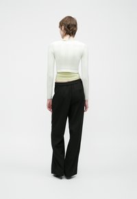 Person med kort brunt hår, iført en hvit kortermet topp med lange ermer, lysegrønn skjorte under, og svarte vide bukser, stående med ryggen til.