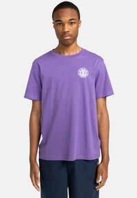 Lilla bomulds T-shirt med rund hals og korte ærmer, med et hvidt logo på venstre brystområde, præsenteret mod en neutral baggrund.