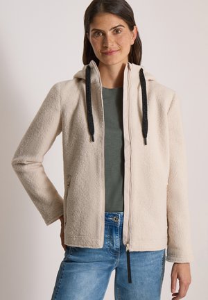 Mujer con cabello castaño que lleva una sudadera beige con cremallera y cordones negros, camisa verde y vaqueros azules sobre un fondo liso.