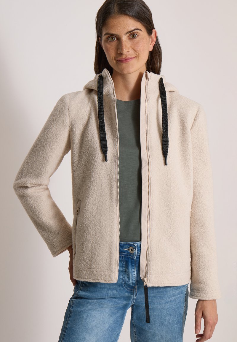 Femme aux cheveux bruns portant un sweat à capuche beige zippé avec des cordons noirs, une chemise verte et un jean bleu sur un fond uni.