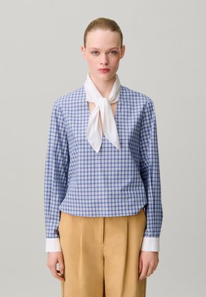 Donna che indossa una camicia a quadri blu a maniche lunghe con polsini bianchi, una sciarpa bianca annodata al collo e pantaloni beige, in piedi davanti a uno sfondo semplice.