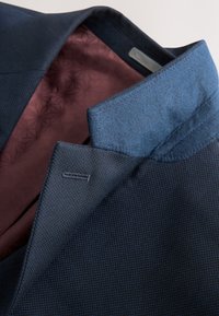 Col de manteau texturé bleu marine avec sous-col en velours bleu foncé et doublure intérieure à motif bordeaux.