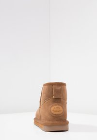 Stivale alla caviglia in suede marrone con punta arrotondata, linguetta sul retro e patch con logo circolare etichettato "EMU Australia" sul tallone. Suola in gomma piatta.