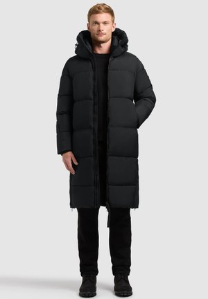 Man draagt een lange zwarte donsjapon met capuchon, zwarte broek en zwarte schoenen, staand tegen een egale lichte achtergrond.