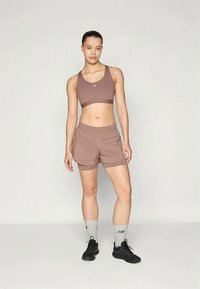 Noor naine seisab, seljas pruun spordibra ja lühikesed püksid, hallid sokid ning mustad spordijalatsid, ühtlase heleda taustaga.