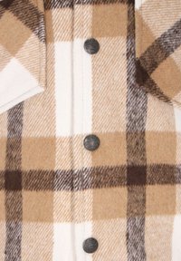 Camisa de franela a cuadros marrón y crema con una textura suave, botones plateados y dos bolsillos en el pecho. Cuenta con un cuello abotonado.