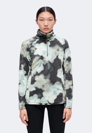 Νέα γυναίκα που κοιτάζει μπροστά, φοράει fleece με φερμουάρ και μακριά μανίκια με μαύρα, γκρι και ανοιχτό μπλε σύννεφο-μοτίβα και μαύρο παντελόνι.