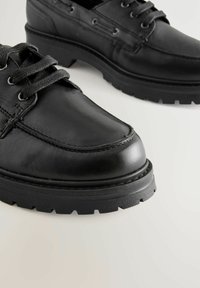 Scarpe in pelle nera con punta arrotondata, finitura testurizzata e suola in gomma spessa con battistrada. Presentano sei asole con lacci neri a contrasto.
