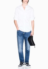 Weißes Langarmhemd, blaue Jeans mit einem leicht ausgewaschenen Finish und einer schwarzen Reißverschlusstasche. Die Schuhe haben schwarz-weiße Streifen.