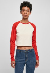 Langärmliges, kurzes Hemd mit einem cremefarbenen Körper und roten Raglanärmeln, aus weichem Baumwollstoff hergestellt. Das Modell kombiniert es mit blauen Jeans.