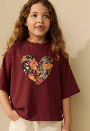Young girl with long wavy hair wearing a maroon shirt with a shiny multicolored sequin heart, beige pants, smiling and looking upward.