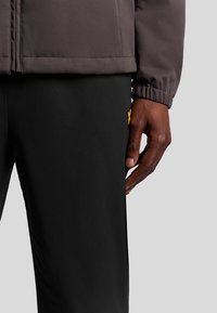 Grijze jacket met een rits en elastische manchetten, gecombineerd met zwarte broek met een klein geel logo. Hand op de zijkant van de broek geplaatst.