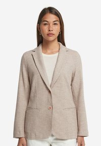 Beige texturerad kavaj med slag, två framfickor och två knappar. Materialet har ett subtilt vävt mönster.