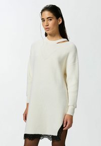 Abito a maglia color crema con texture a coste, presenta un piccolo taglio sulla spalla e un dettaglio in pizzo nero all'orlo, maniche lunghe e vestibilità rilassata.