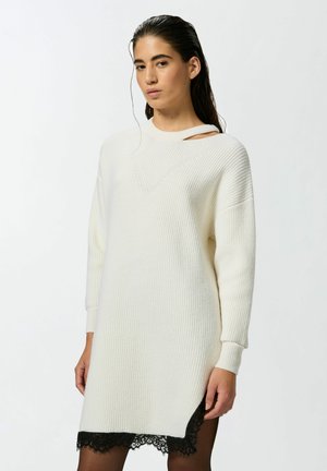 Abito a maglia color crema con texture a coste, presenta un piccolo taglio sulla spalla e un dettaglio in pizzo nero all'orlo, maniche lunghe e vestibilità rilassata.
