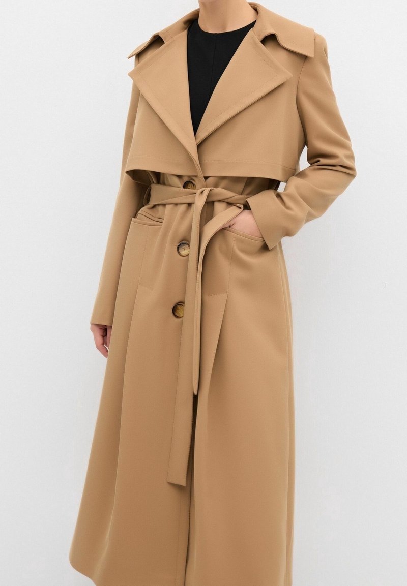 Femme portant un long trench-coat camel avec de grands revers, une ceinture nouée à la taille et deux boutons visibles à l'avant.