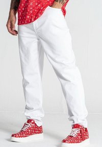 Pantaloni bianchi con una vestibilità rilassata, abbinati a sneakers rosse e bianche con un motivo. L'outfit presenta una vivace camicia rossa a motivi.