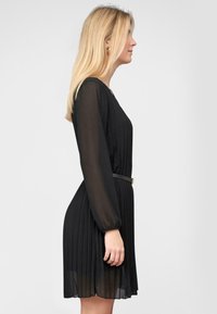 Černé plisované šaty s dlouhými průhlednými rukávy, přiléhavý pas zvýrazněný úzkým páskem a délka po kolena. Hladká textura a elegantní design.