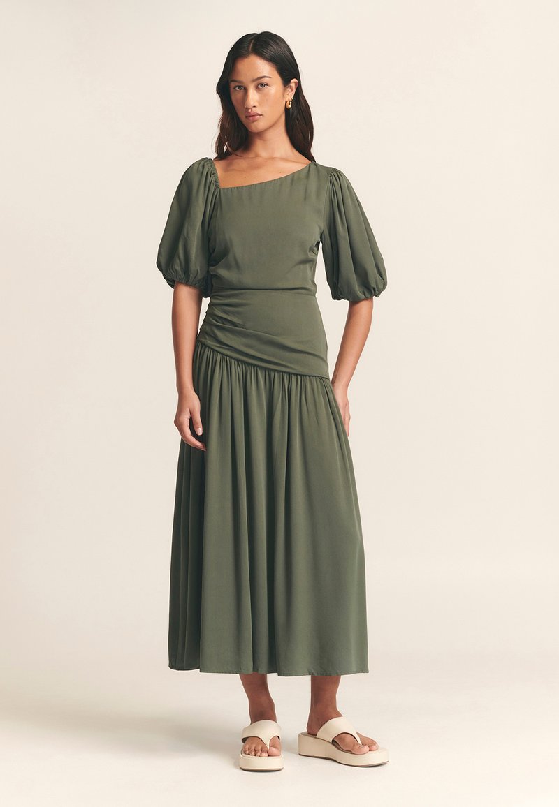 Vestito verde oliva con maniche a sbuffo, scollatura quadrata e vita arricciata. Gonna fluida che arriva a metà polpaccio. Abbinato a sandali platform color crema.