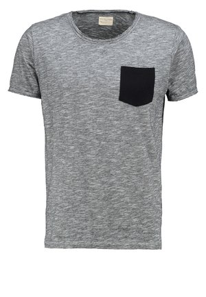 Mand iført grå t-shirt med sort lomme, sorte bukser, hvide sneakers med mønstre, holder sort læderrygsæk ved siden af.