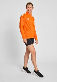 Herre orange atletisk jakke med frontlynlås, sidelommer og hvide logoaccents, parret med sorte shorts og sorte sportsko.