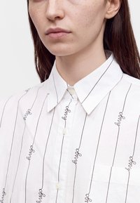 Jeune personne aux cheveux lisses et foncés portant une chemise blanche boutonnée avec de fines rayures verticales noires et le mot "hugo" imprimé à plusieurs reprises.