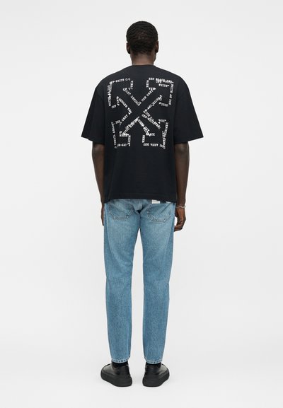 Άνδρας που φοράει μαύρο φαρδύ μπλουζάκι με λευκό αφηρημένο σχέδιο κειμένου στην πλάτη, ανοιχτό μπλε τζιν και μαύρα παπούτσια, στέκεται με την πλάτη προς τα πίσω.