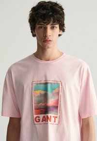 Lichtroze katoenen t-shirt met een centraal grafisch ontwerp van een kleurrijk landschap en het woord "GANT" in dikke rode letters.