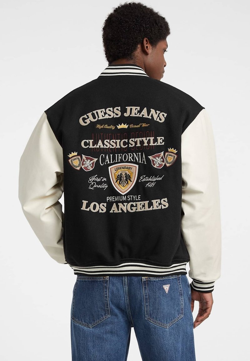 Mandens sorte og creme varsity jakke med ribbede manchetter, der har broderede logoer og tekst på ryggen, denim jeans er synlige nedenfor.