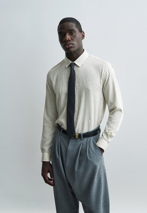 Muž v bielej košeli s tenkými pruhmi na dlhý rukáv, čiernou kravatou, sivými plisovanými nohavicami a čiernym opaskom, stojaci s jednou rukou v vrecku.