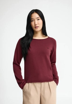 Ung kvinde med langt sort hår iført en bordeaux langærmet sweater og beige bukser, stående med hænderne i lommerne mod en hvid baggrund.