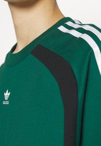 Grön Adidas-mössa med vita tre ränder på axlarna, svarta paneler och Adidas trefoil-logotypen på bröstet.
