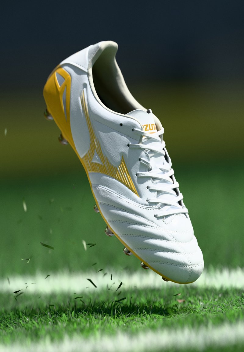 Λευκό και κίτρινο ποδοσφαιρικό παπούτσι Mizuno σηκώνοντας χόρτο σε πράσινο γήπεδο με λευκές γραμμές ορίων.