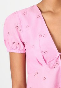Rosa bluse med puffede ærmer, prydet med blomsterbroderi og små udskæringer. Binder ved halsen, lavet af letvægtsstof.