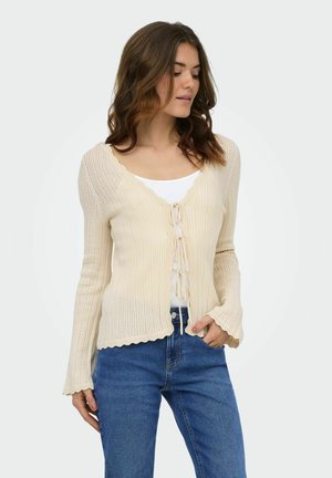 Femme aux cheveux bruns portant un cardigan côtelé crème noué devant sur un top blanc et un jean bleu, debout avec une main dans la poche.