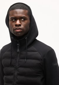 Άνδρας που φοράει μαύρο μπουφάν με κουκούλα και καπιτονέ μπροστινό μέρος, κοιτάζοντας προς τα πλάγια μπροστά σε απλό φωτεινό φόντο.