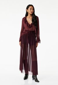 Burgundy läpinäkyvä maksi mekko, jossa on röyhelökaula-aukko, pitkät hihat ja nyöritettävä vyötärö; kerrosmainen rakenne, yhdistettynä mustiin nilkkasandaaleihin.